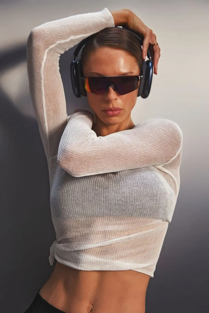 Jules in a sheer white knit top and headphones, one arm raised over her head